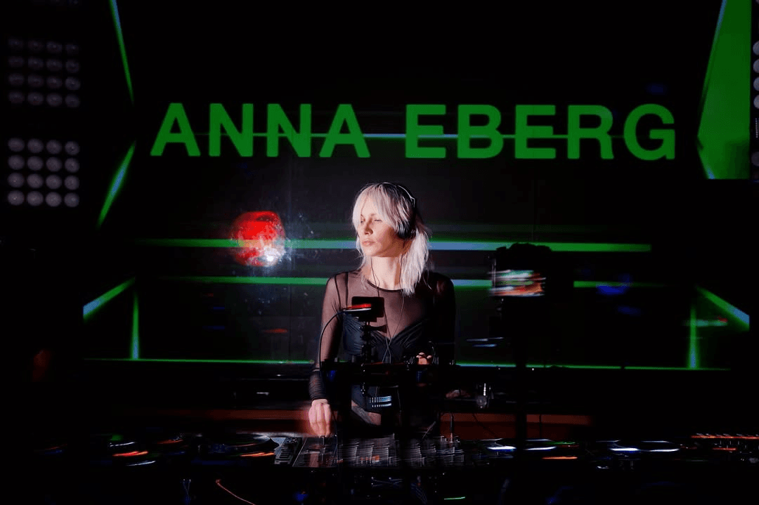 Blonde DJ Anna Eberg wearing headphones performs at a booth with vibrant green neon background text.